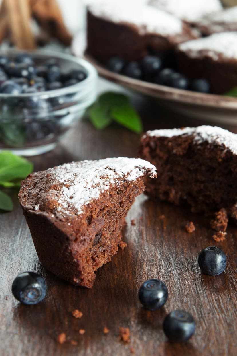 Moist chocolate cakes dusted with sugar and topped with blueberries rest on a dark rustic wooden surface.