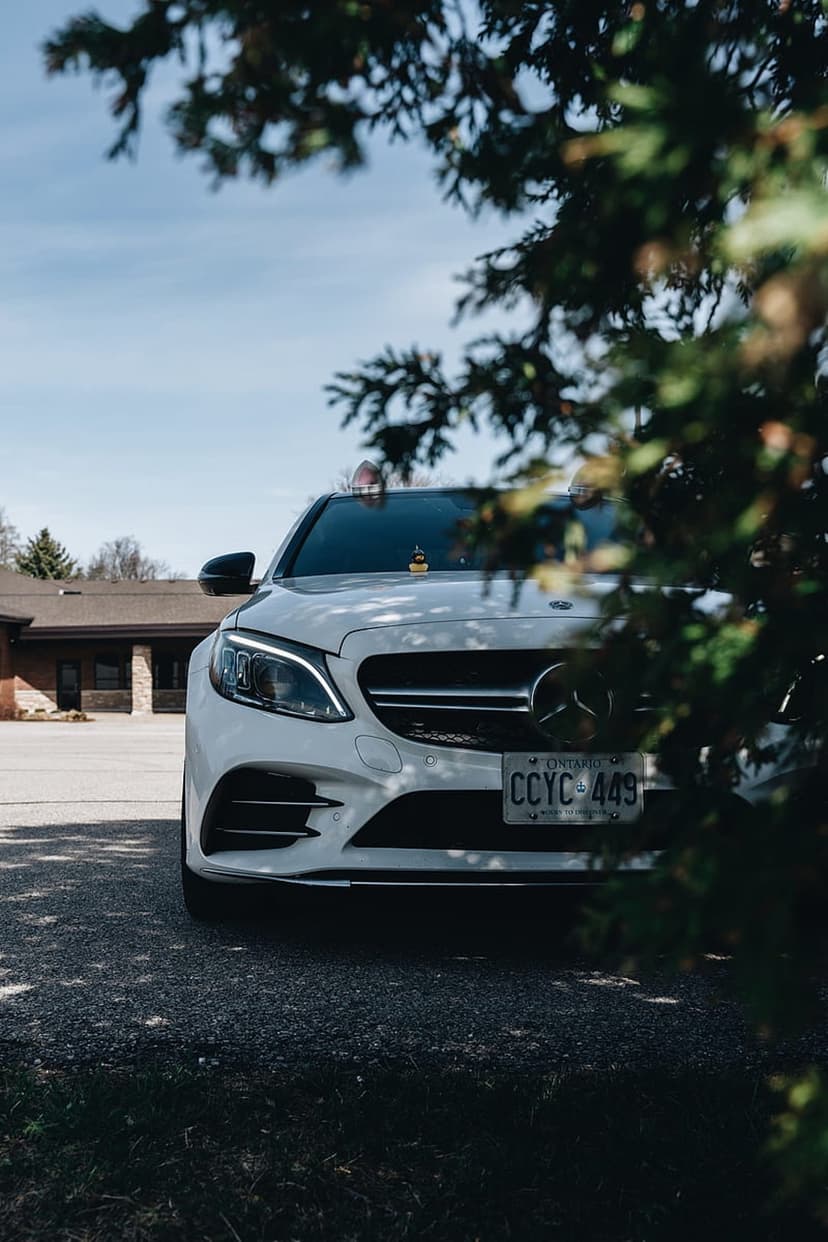 White Mercedes-Benz C-Class sedan parked on gravel with headlights glowing under green tree branches in daylight.