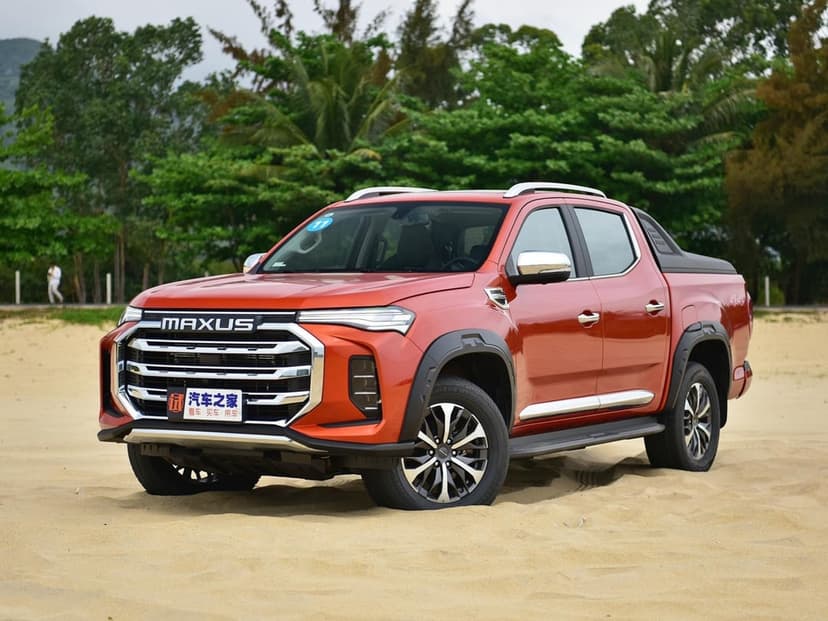 Orange Maxus pickup truck with a large chrome grille parked on a sandy beach against lush green trees.