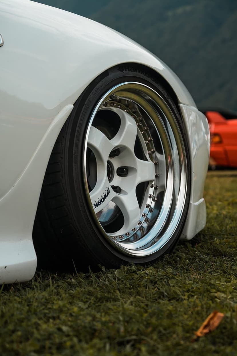 White multi-spoke custom wheel with polished rim on green grass against a blurred red car background.