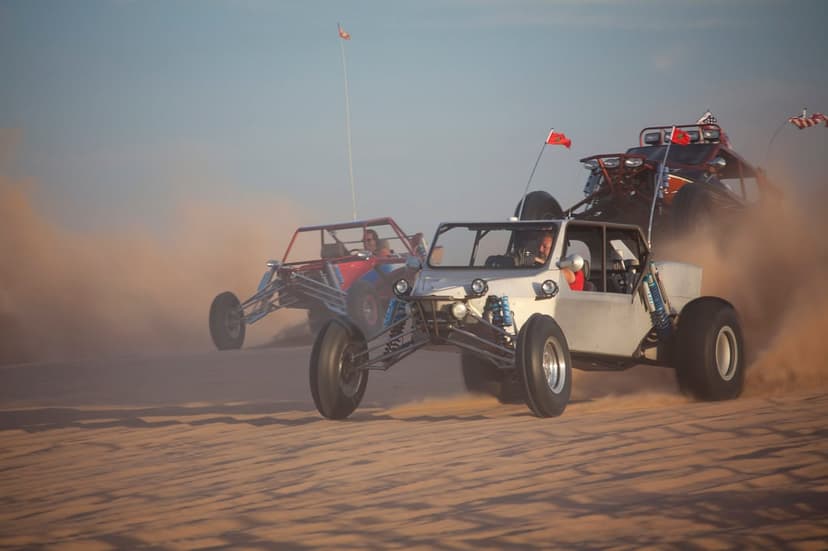 Two off-road dune buggies race across desert sands while kicking up massive dust clouds under a clear blue sky.