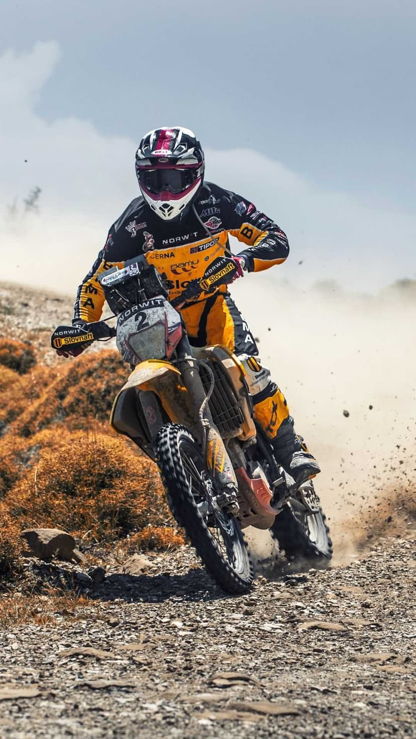 Dirt bike racer in yellow gear leans into a turn on a rocky trail while clouds of dust rise from the rear tire.