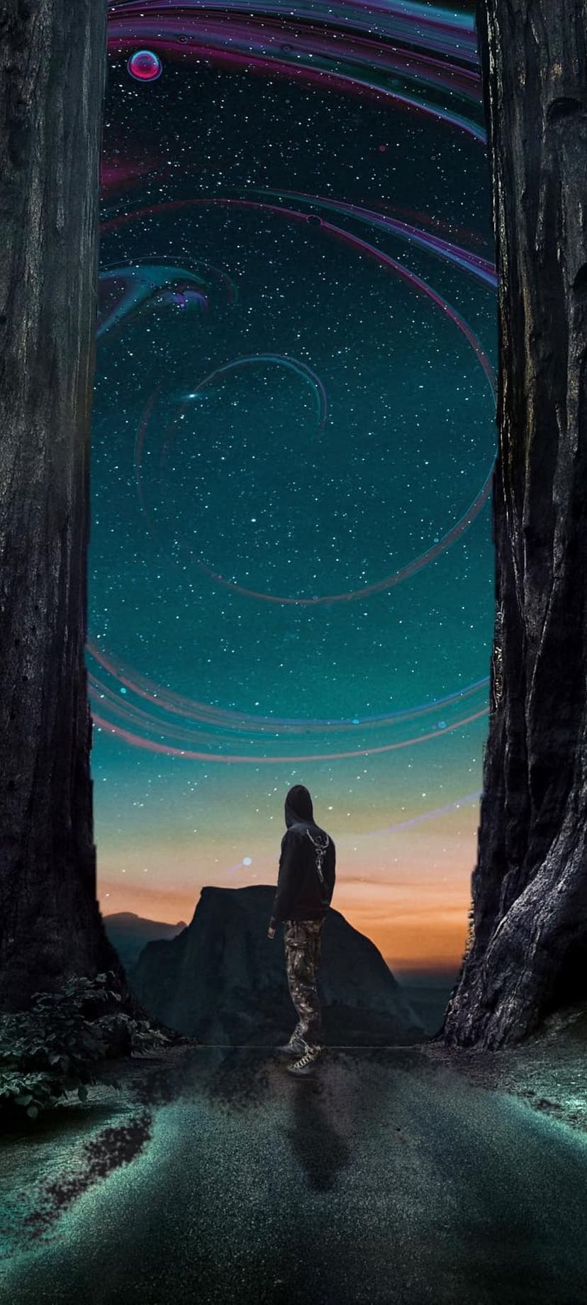 Person in a hoodie stands between dark trees looking at a swirling blue and purple galaxy over mountains.