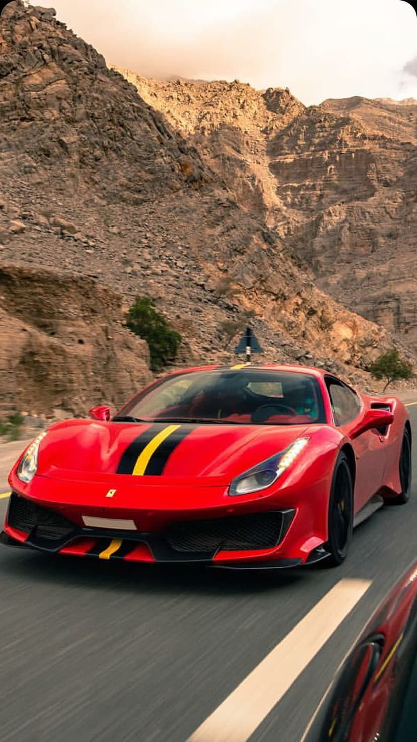 Red Ferrari with racing stripes speeds along a winding mountain road between sun-drenched rocky cliffs.