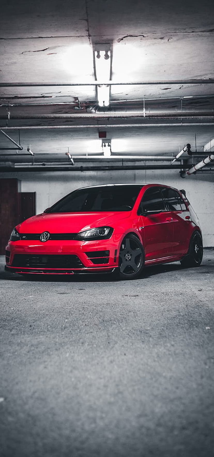 Red Volkswagen Golf R hatchback with black wheels parked in a dark concrete garage under industrial lights.