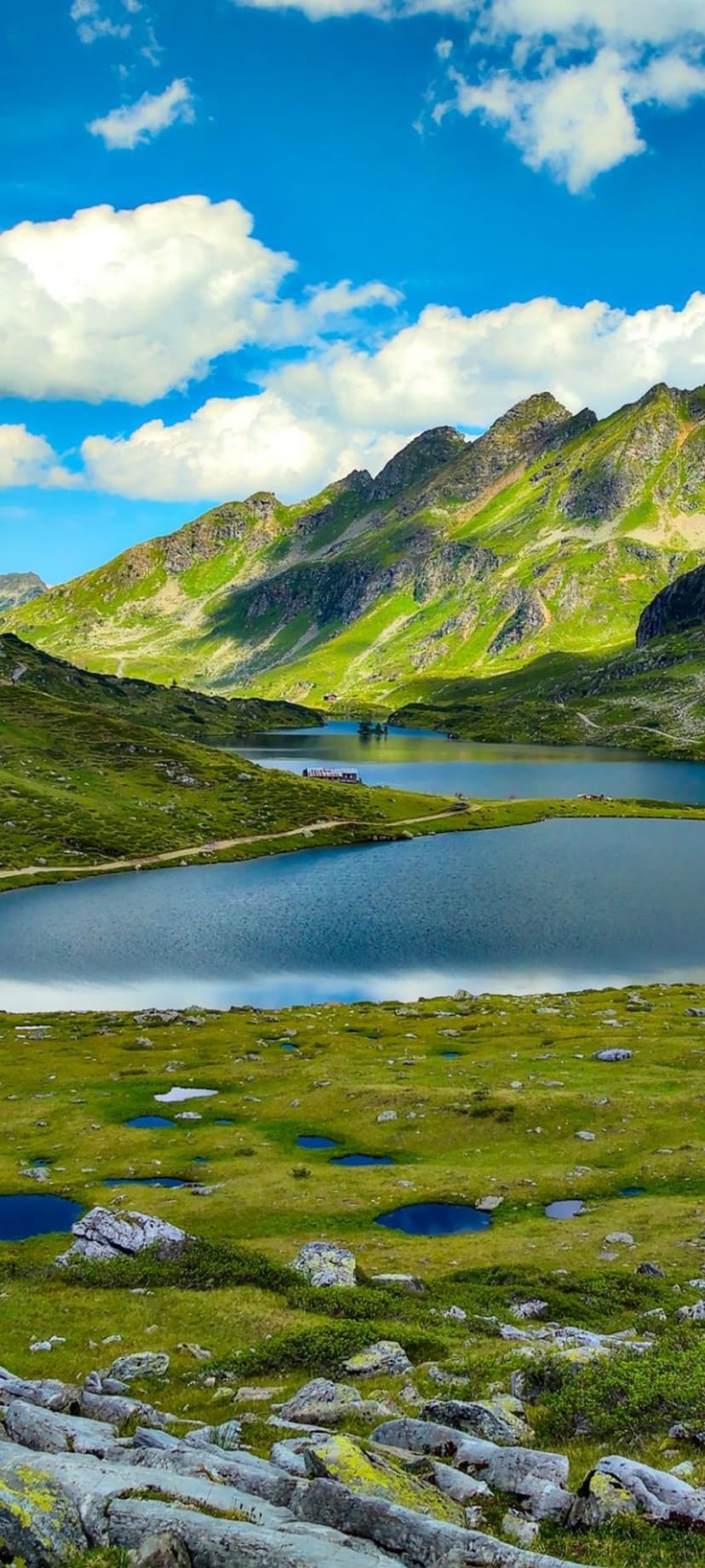 Steep green mountains surround a clear alpine lake under a blue sky with white clouds and rocky foreground details.