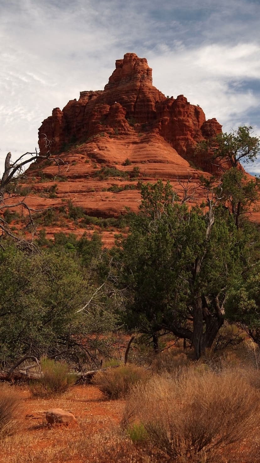 Towering red sandstone formation rises above green shrubs and desert soil under a cloudy sky in Sedona.