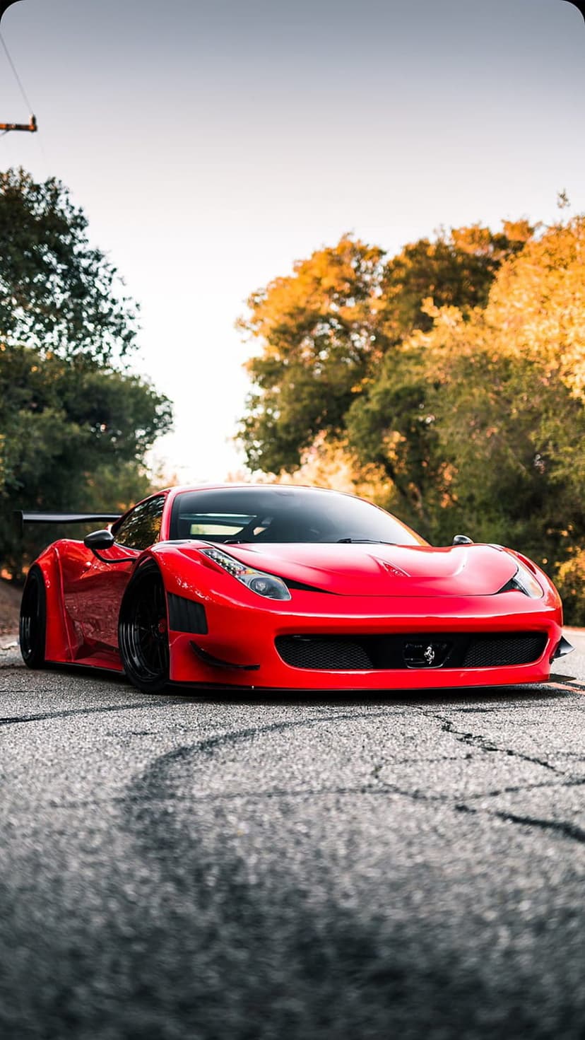 Red Ferrari 458 Italia with a wide body kit and racing spoiler on an asphalt road lined with autumn trees.