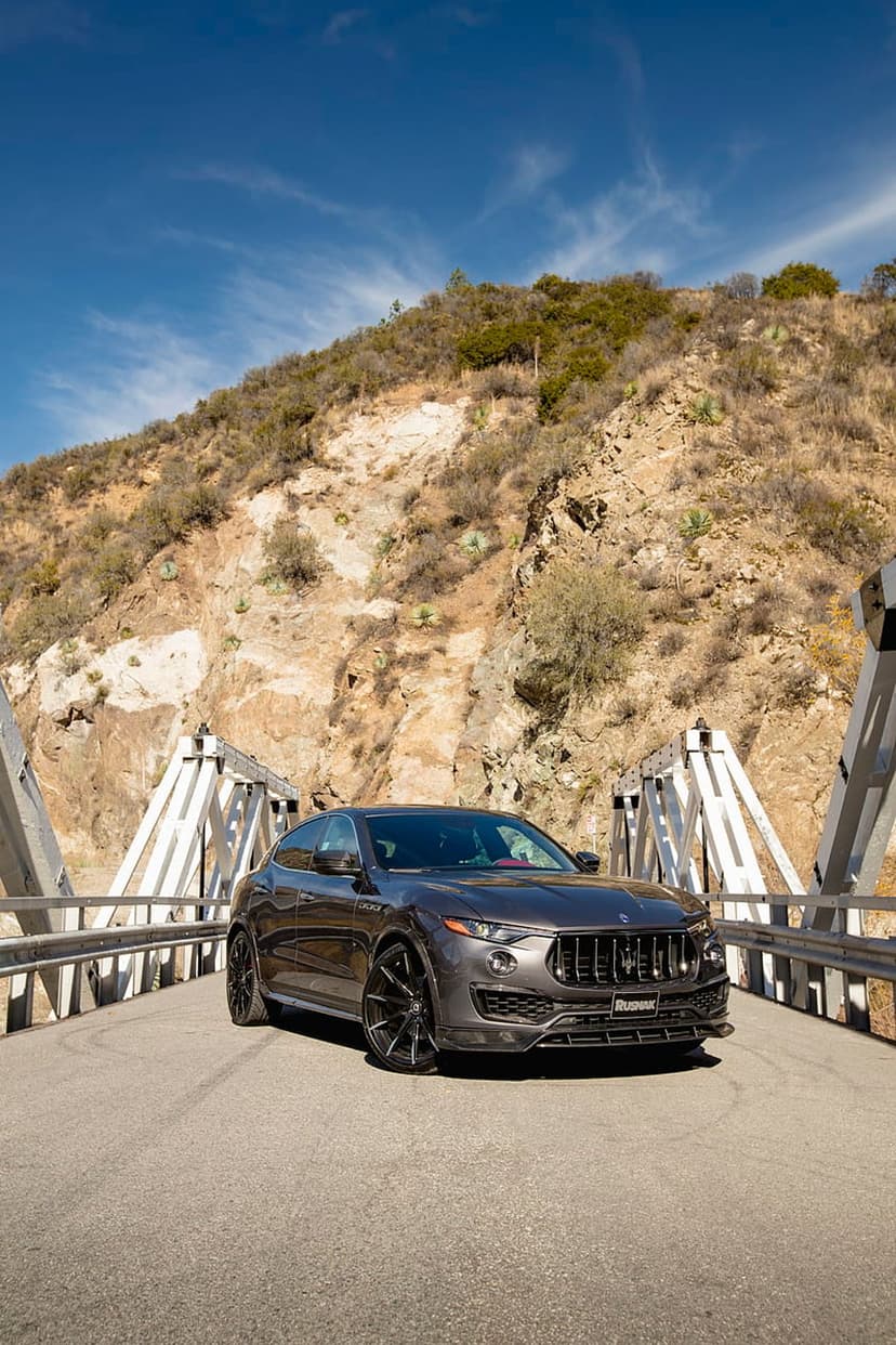 Maserati Levante SUV parked on a white metal bridge with a dry mountain range under a clear blue sky.