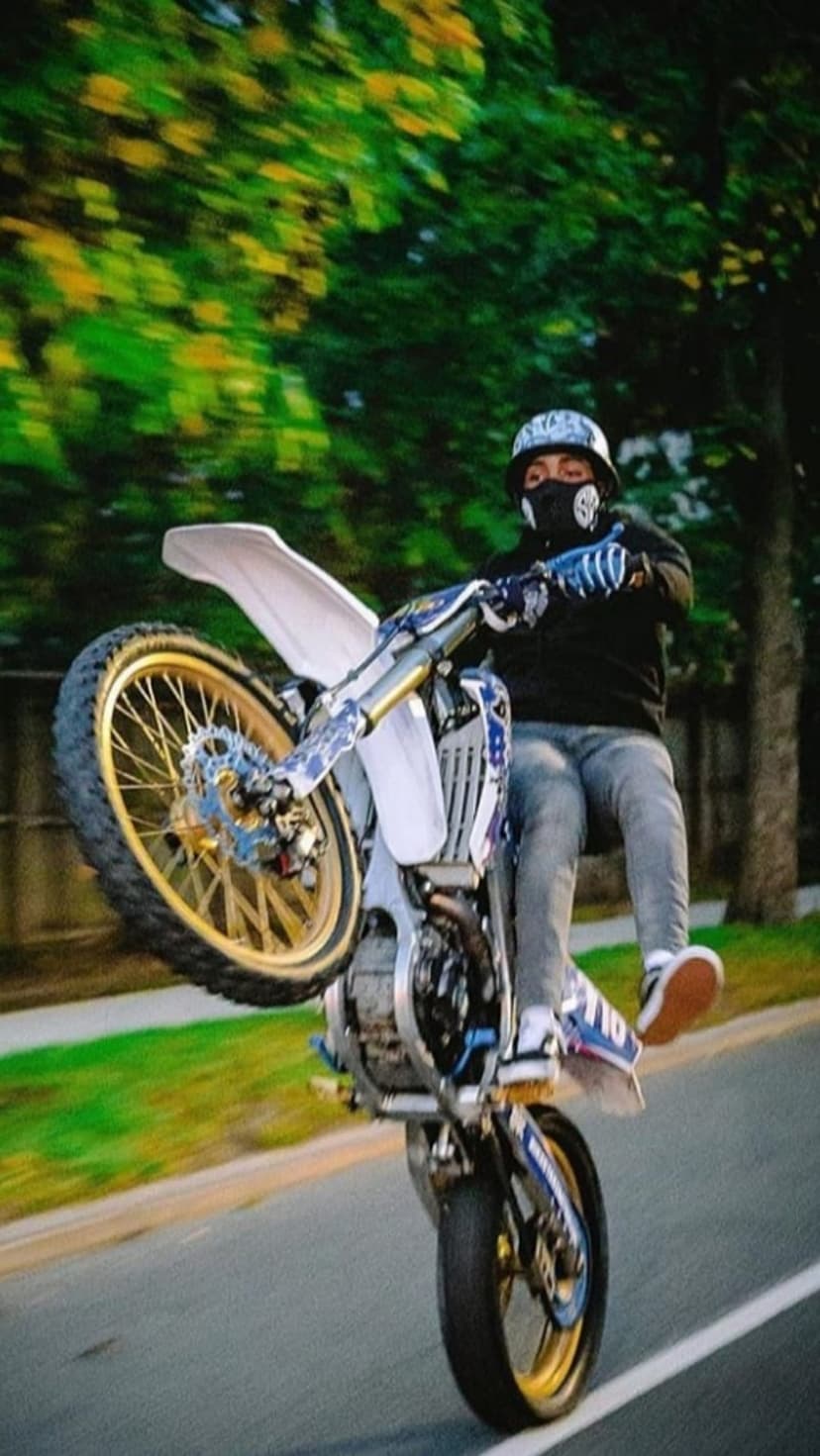 Motocross rider in a black hoodie performs a wheelie on a street with gold-rimmed wheels and blurred trees.