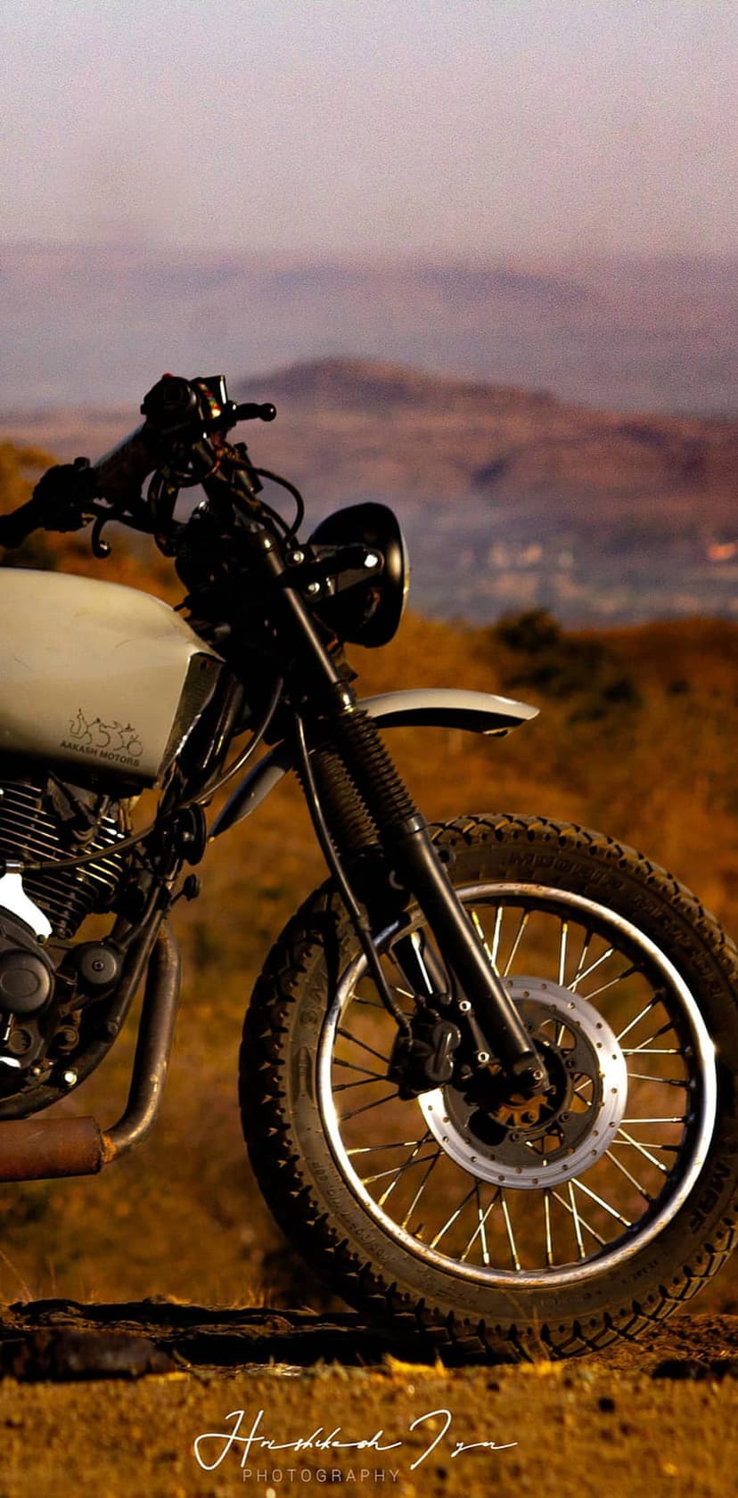 Vintage scrambler motorcycle parked on a dry grassy hill against a hazy sunset sky with rolling hills behind it.