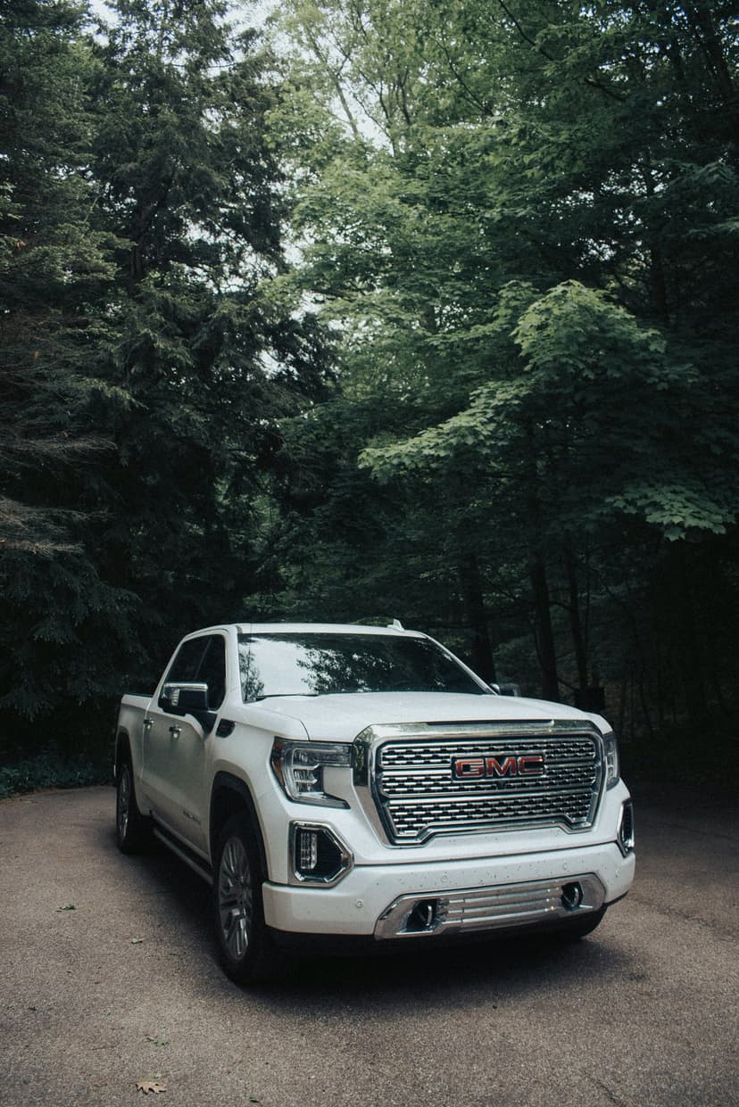 White GMC Sierra pickup truck on a gravel road in a lush green forest with sunlight filtering through tall trees.
