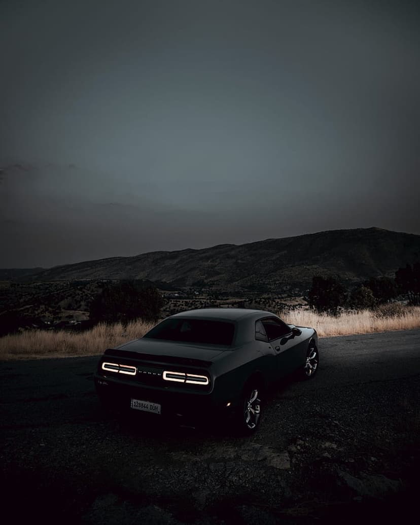 Black Dodge Challenger with glowing red taillights parked on a dirt mountain road during a purple twilight dusk.