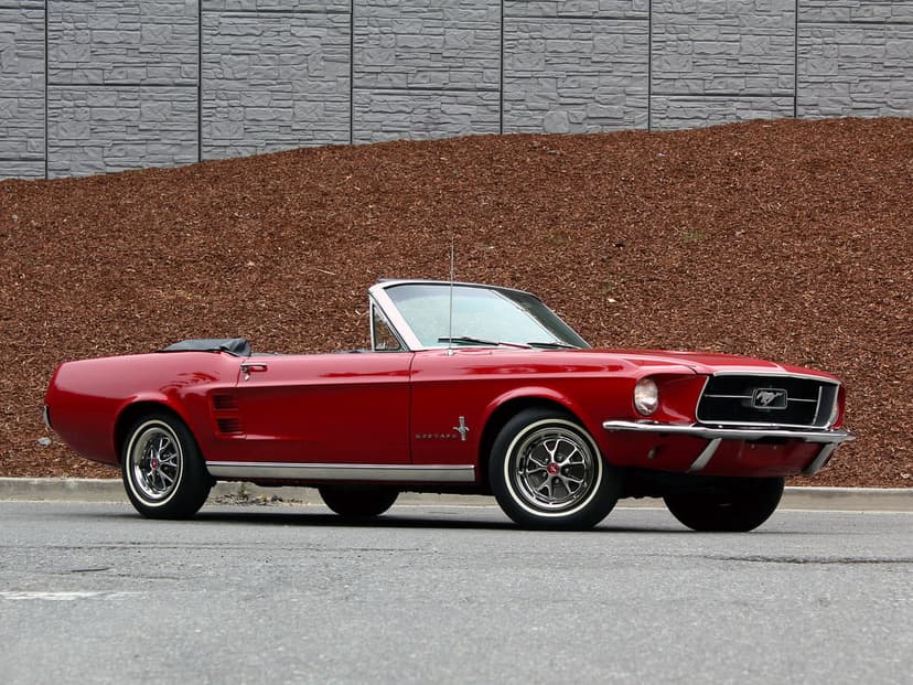 Red 1967 Ford Mustang convertible parked on gray asphalt against a textured brown mulch hill and stone wall.