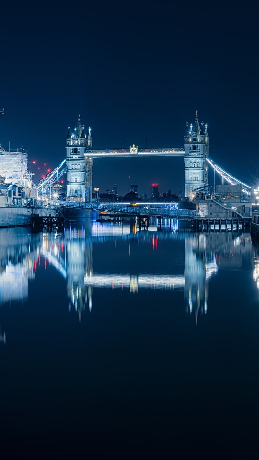 Tower Bridge illuminated with blue lights at night reflects on the River Thames with dark skies in the background.
