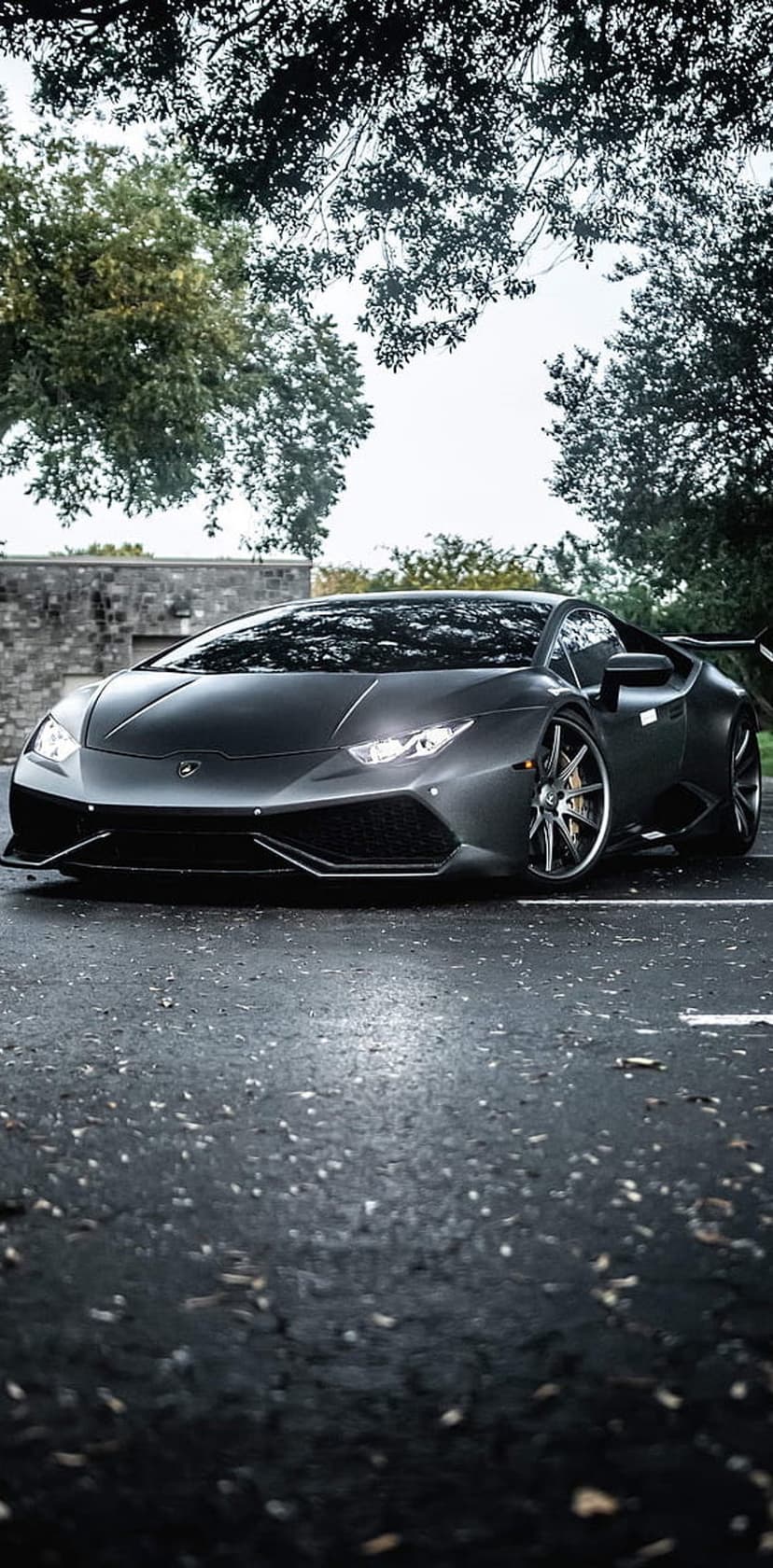 Matte black Lamborghini Huracan parked on a wet pebbled road with glowing headlights and a stone wall backdrop.