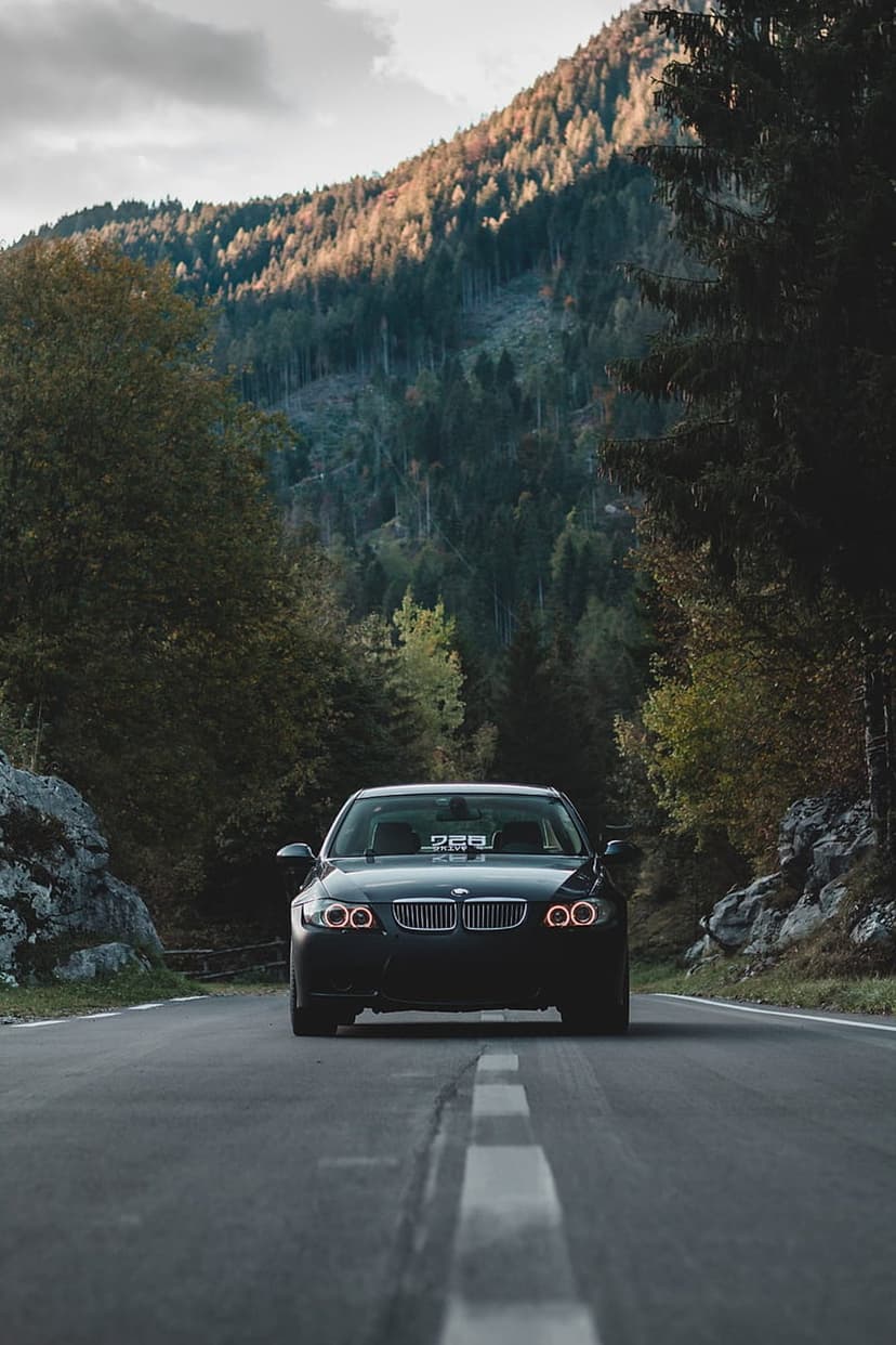 Black BMW sedan drives on a winding mountain road surrounded by golden autumn forests under a cloudy sunset sky.