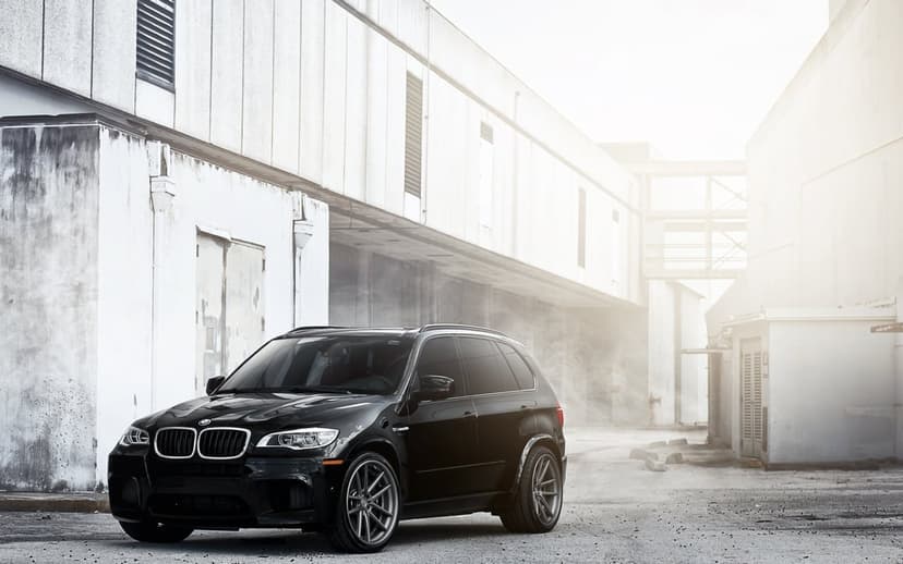 Black BMW X5 M SUV parked between tall white industrial buildings with soft sunlight and a misty background.