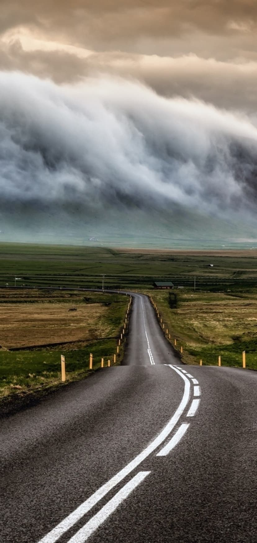 Asphalt road curves through golden and green fields toward distant hills under a heavy, dark overcast sky.
