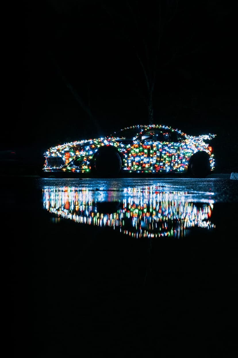 Supercar draped in multi-colored holiday lights with a shimmering reflection on wet pavement at night.
