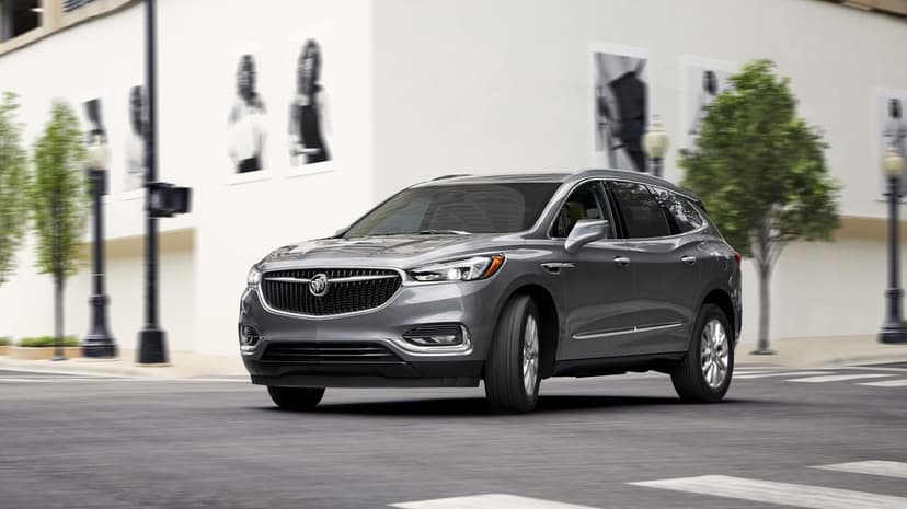 Grey luxury SUV driving through a city crosswalk past white buildings and green trees with motion blur.