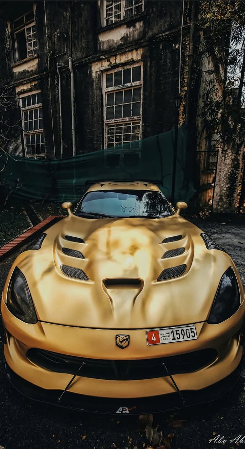 Golden Dodge Viper supercar reflects light before a weathered brick building with surrounding green foliage.