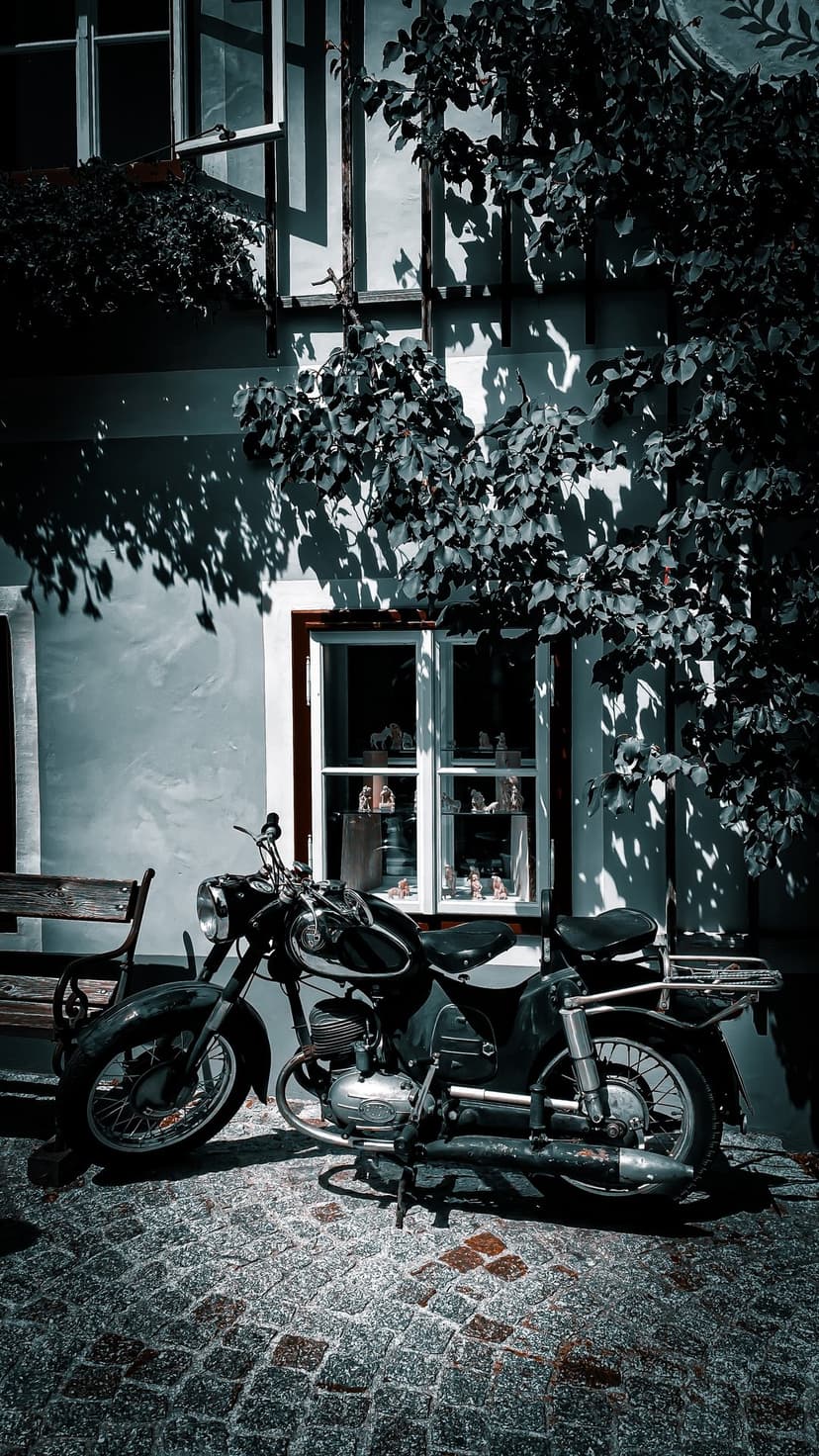 Vintage black motorcycle on a cobblestone path beside an ivy-covered building with dappled sunlight shadows.