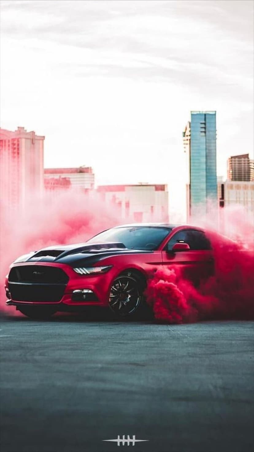 Red Mustang car drifting with thick pink smoke clouds against a city skyline and cloudy gray sky background.