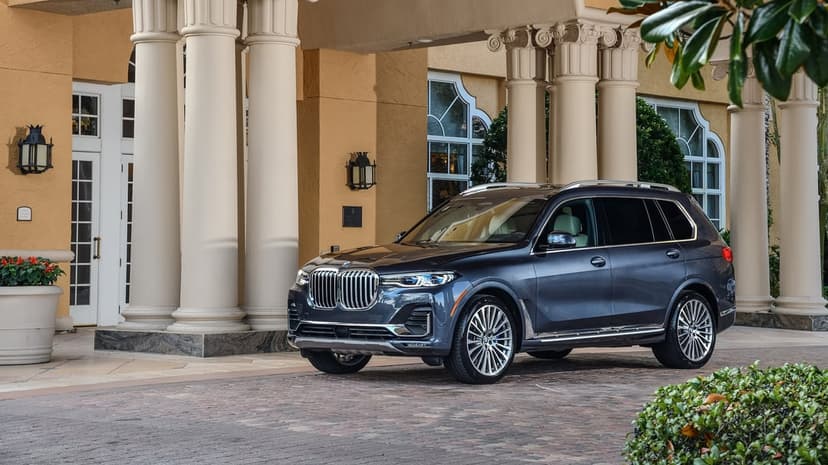 A dark grey BMW X7 luxury SUV parked in front of a grand building with massive beige columns and green leaves.