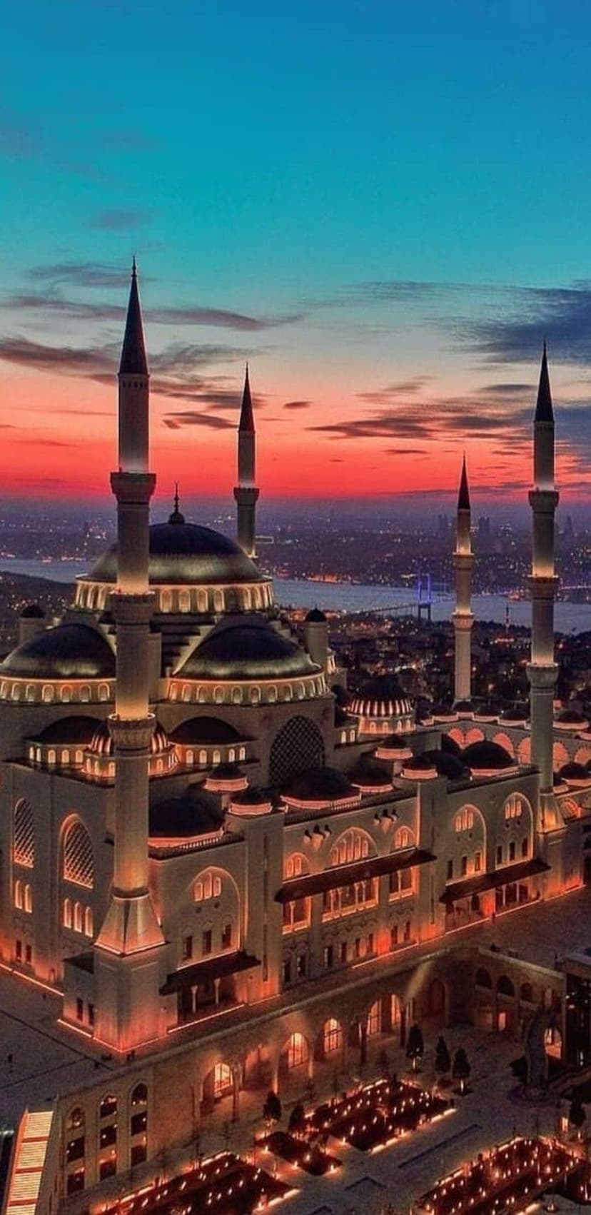 Illuminated mosque domes and minarets stand against a red and blue sunset sky with distant city lights.