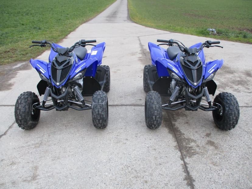Two blue Yamaha Raptor ATVs parked side-by-side on a narrow concrete path between lush green grass fields.