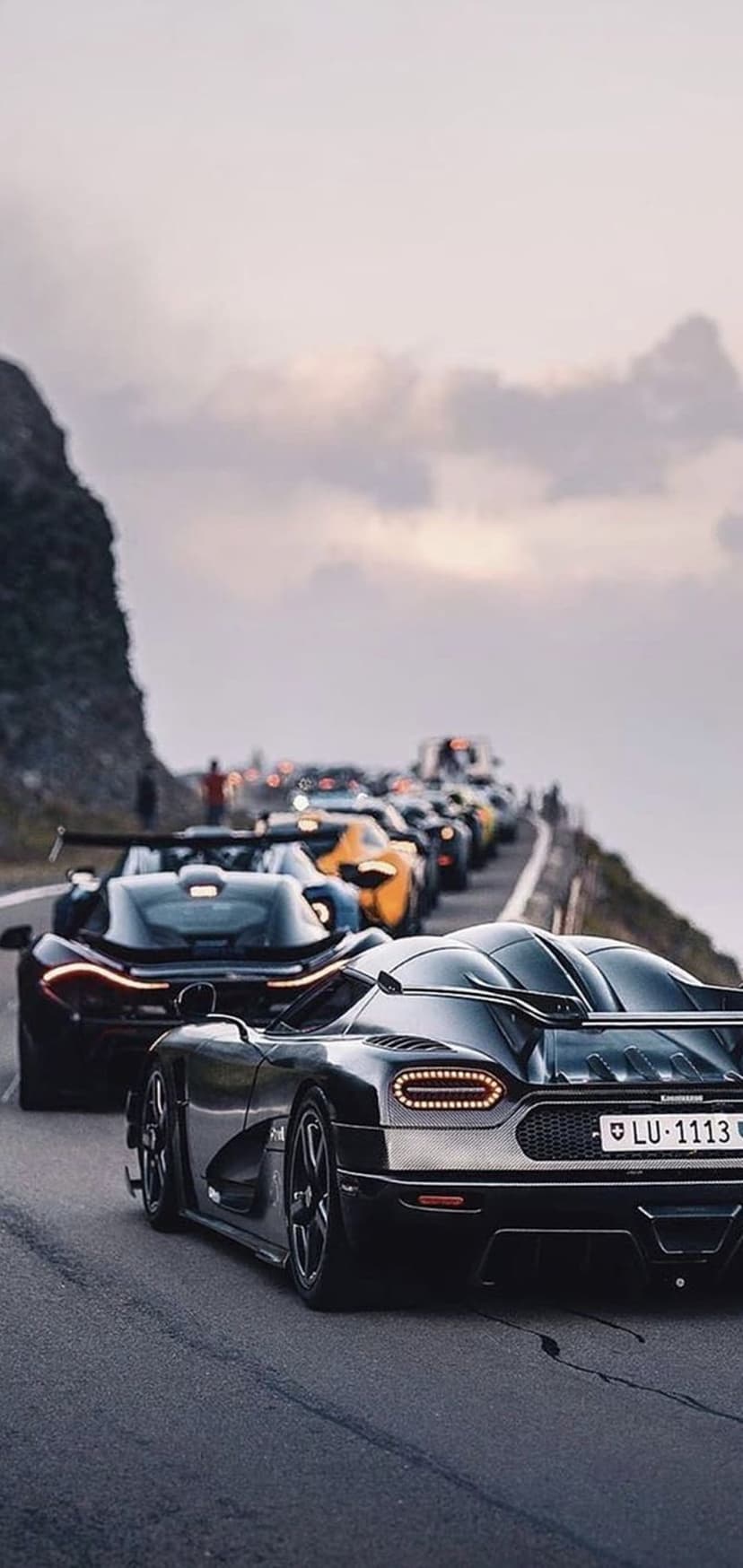 Koenigsegg and McLaren P1 lead a supercar convoy on a winding mountain road beneath dark storm clouds.