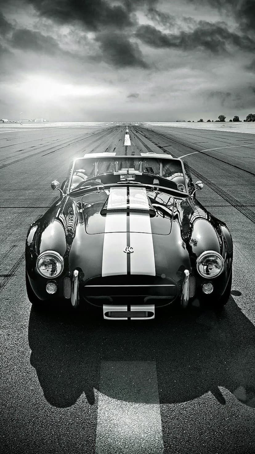 Shelby Cobra with racing stripes rests on a dark runway under a cloudy sky in a high-contrast monochrome scene.