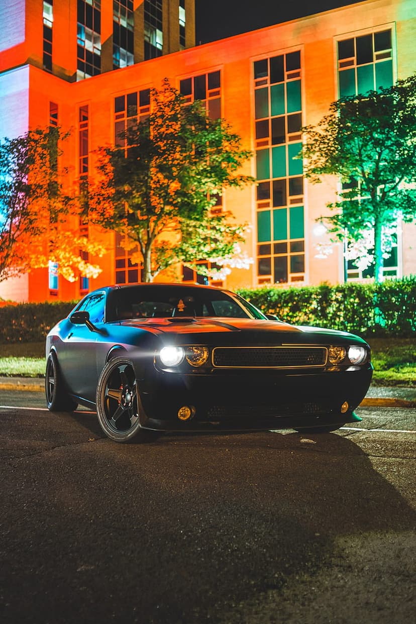 Black Dodge Challenger parked at night under orange and green building lights with glowing headlights on asphalt.