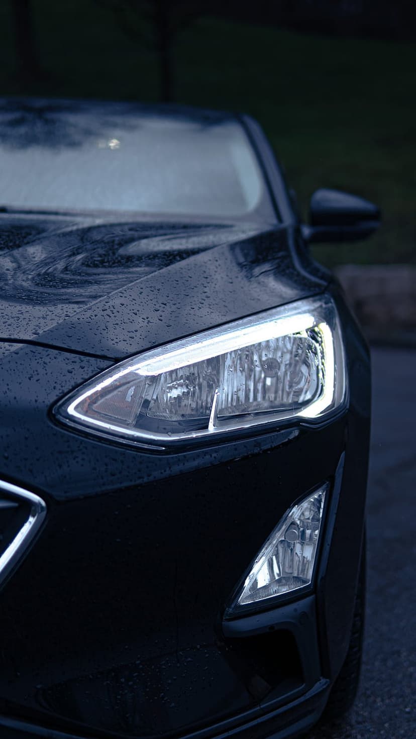 Car headlight glows through falling rain on a dark metallic surface with visible water droplets and reflections.