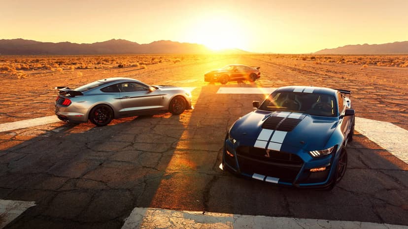 Blue Shelby GT500 with white stripes and silver Mustang on a desert runway at sunset under a golden sky.