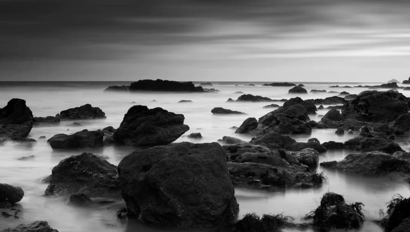 Dark craggy rocks stand against a misty white tide under a streaked gray sky in a vertical coastal scene.