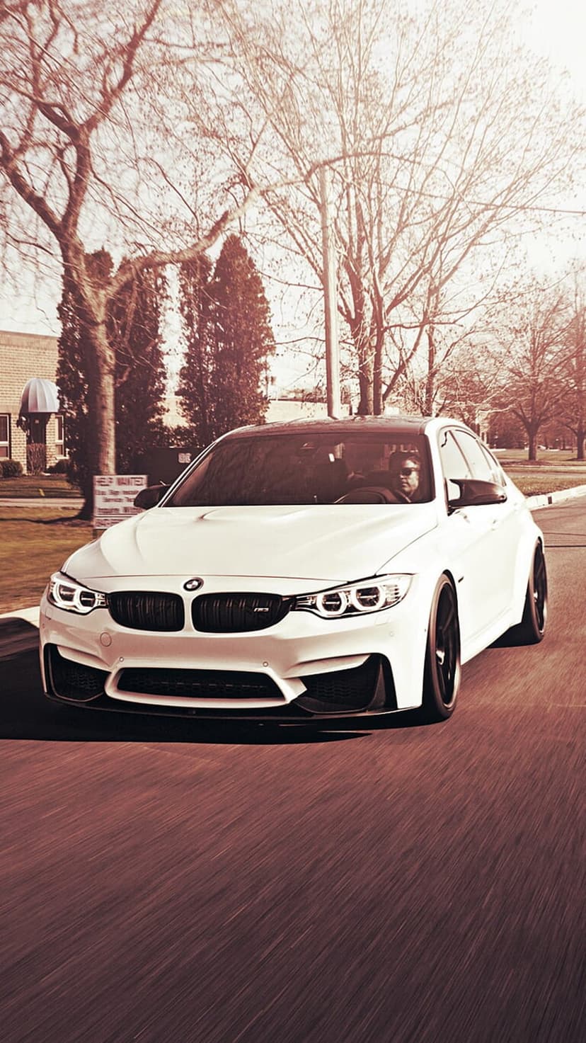 White BMW M4 driving on a suburban road with motion blur and sunlight reflecting off the front grille.