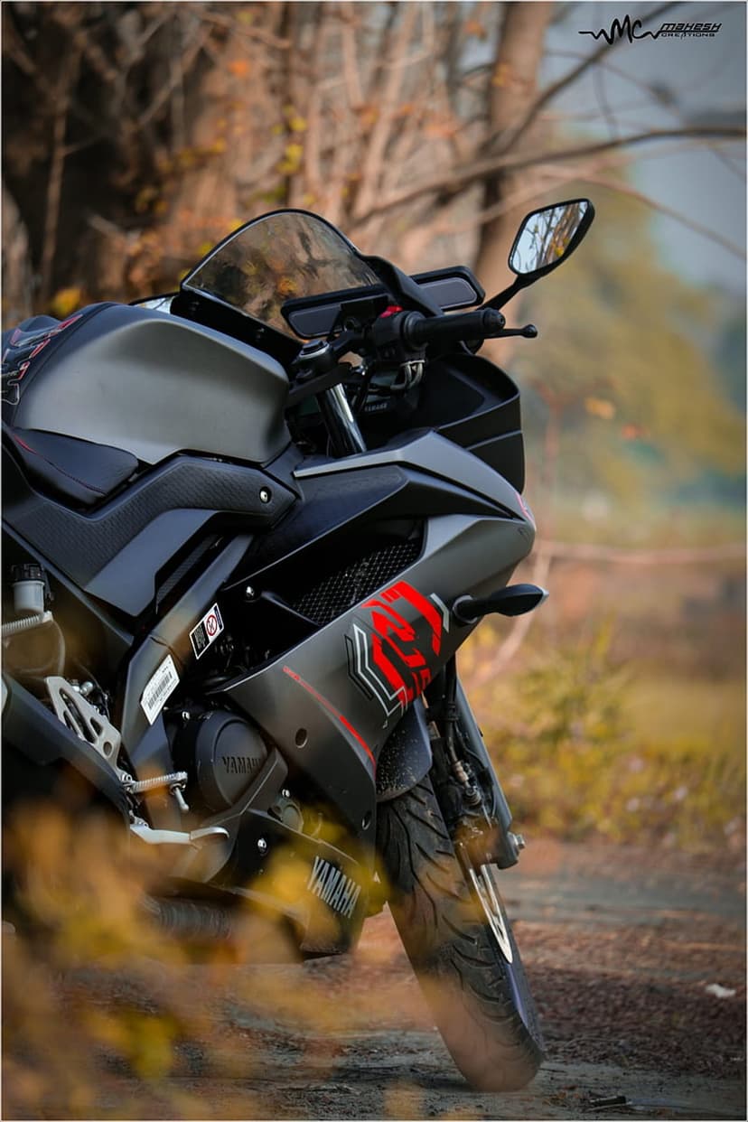 Dark Yamaha R15 sportbike with red decals parked in an autumn forest with blurred orange foliage background.
