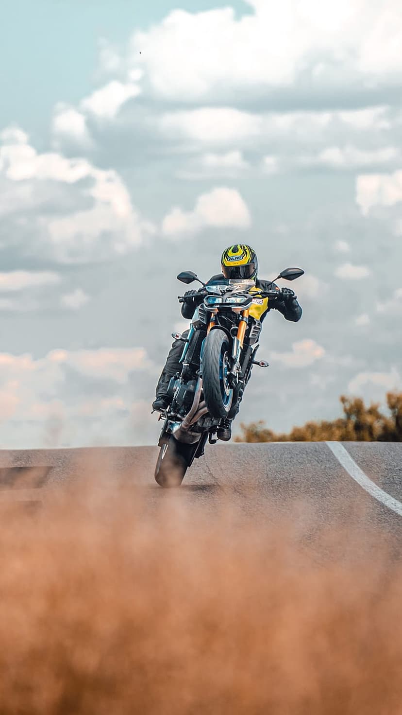 Motorcyclist in a yellow helmet performs a wheelie on a dusty road under a bright blue sky with white clouds.