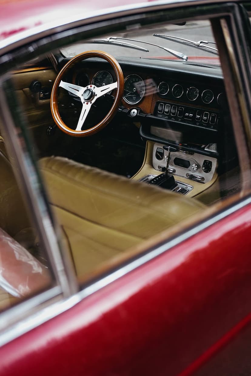 Wood steering wheel and tan leather seats inside a vintage red car with analog dashboard gauges and chrome details.