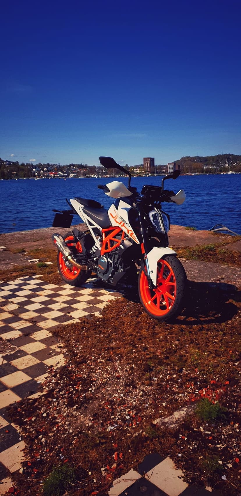 KTM Duke motorcycle in orange and white parked on a rocky shore beside a bright blue lake and distant hills.