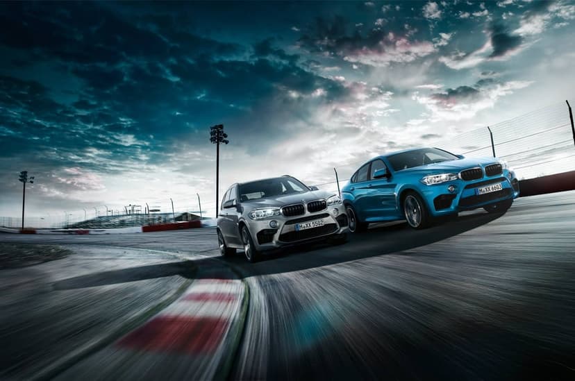 Two BMW SUVs speeding on a race track with motion blur, red rumble strips, and a dramatic cloudy sky.
