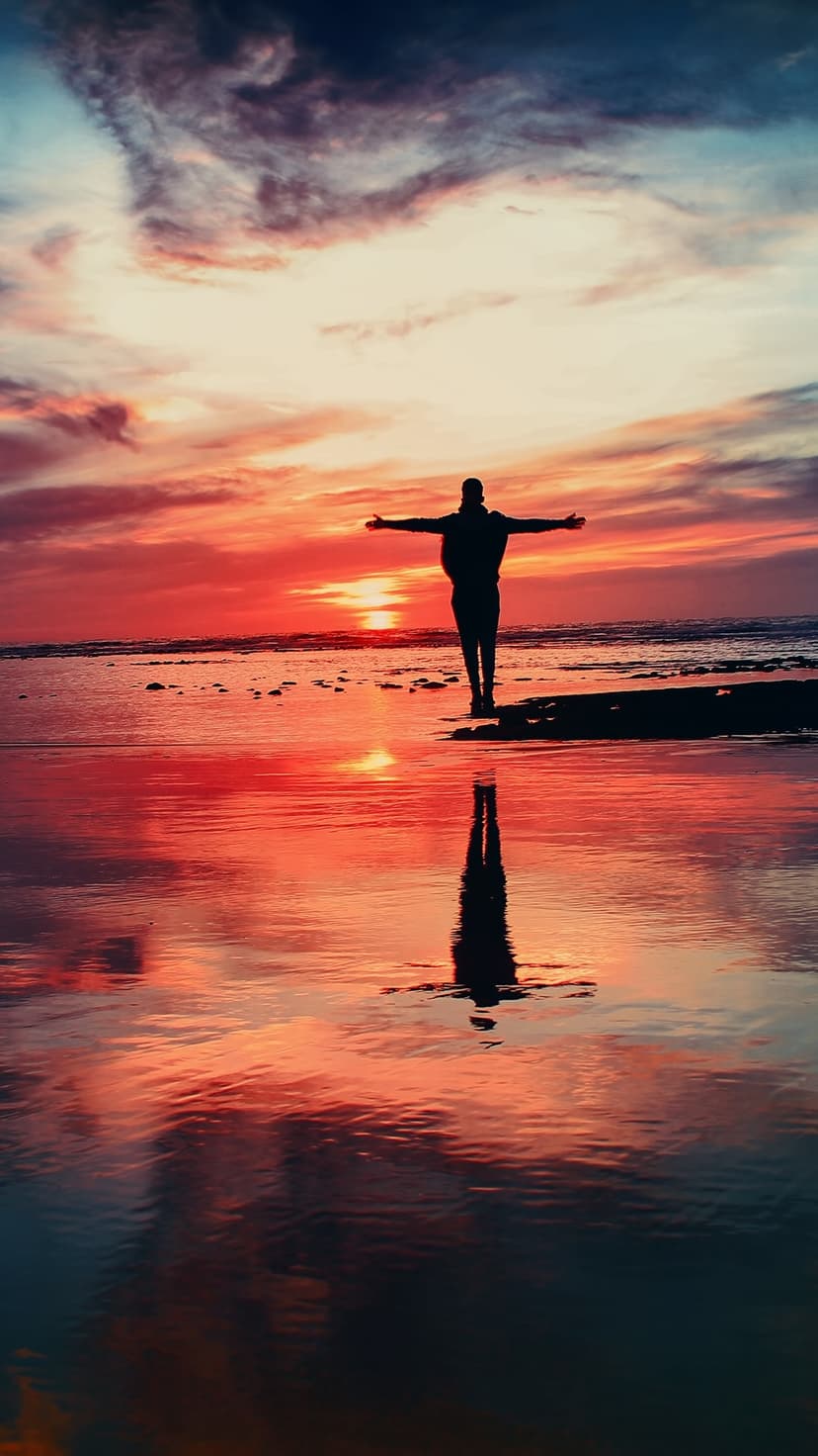 Lone person silhouette with arms wide on a wet beach reflecting a red and purple sunset sky on the water.
