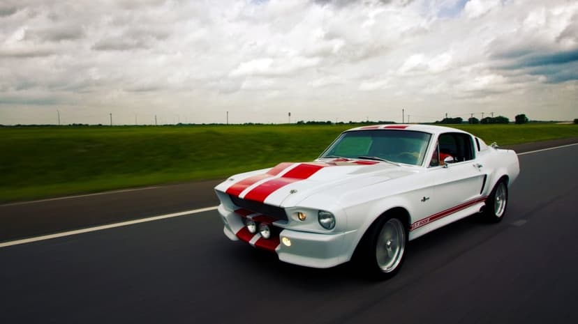 White Shelby GT500 with red stripes speeding on an open highway under a dramatic cloudy sky with green fields.