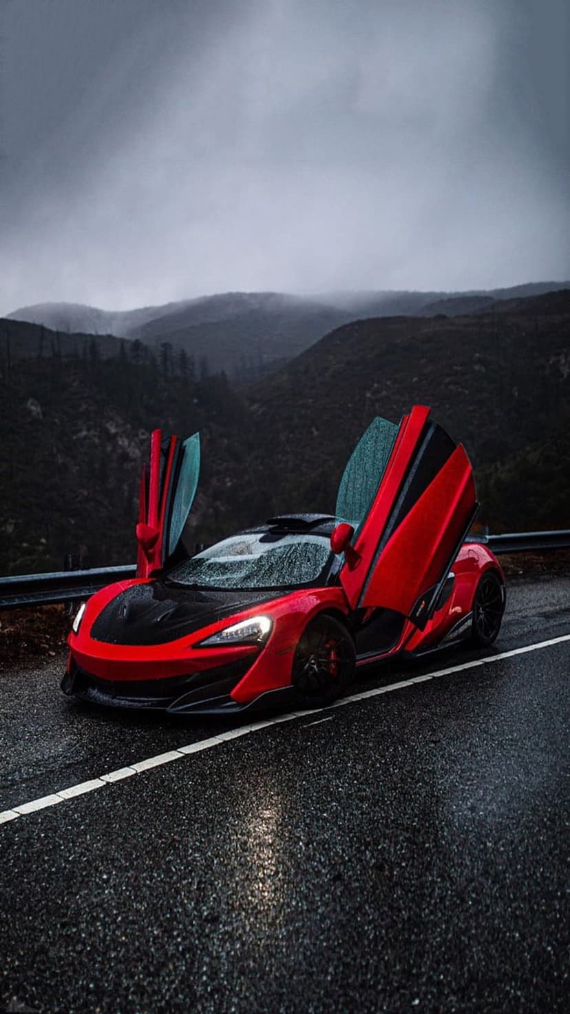 Red McLaren supercar with open scissor doors parked on a wet mountain road surrounded by mist and grey clouds.