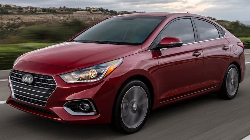 Red Hyundai sedan driving on an open road with motion blurred wheels and a hilly background under grey skies.