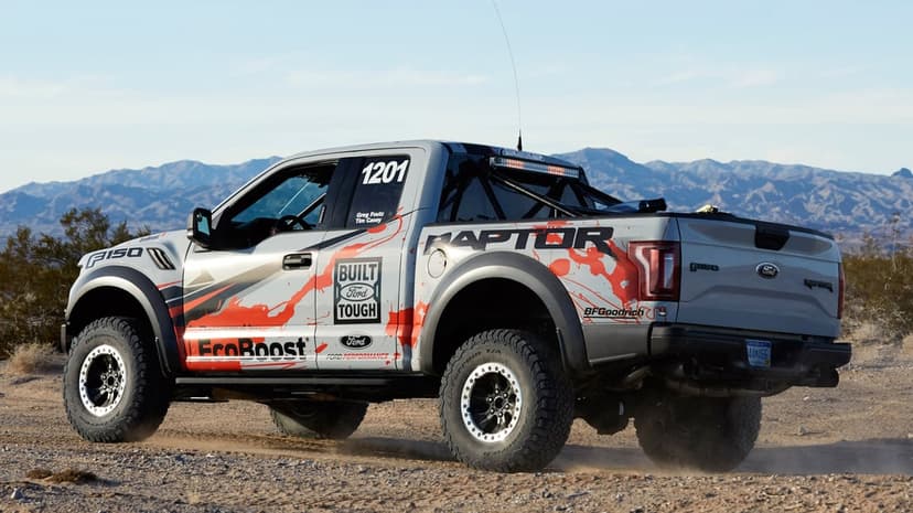 Silver Ford F-150 Raptor race truck with red graphics driving through a dusty desert landscape under a clear sky.