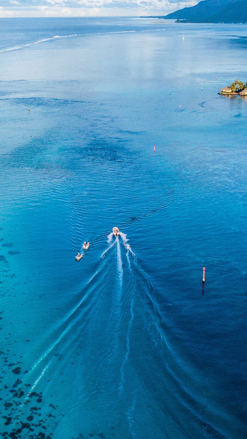 A small boat pulls two tubers across a vast turquoise ocean, leaving long white wakes on the calm water surface.