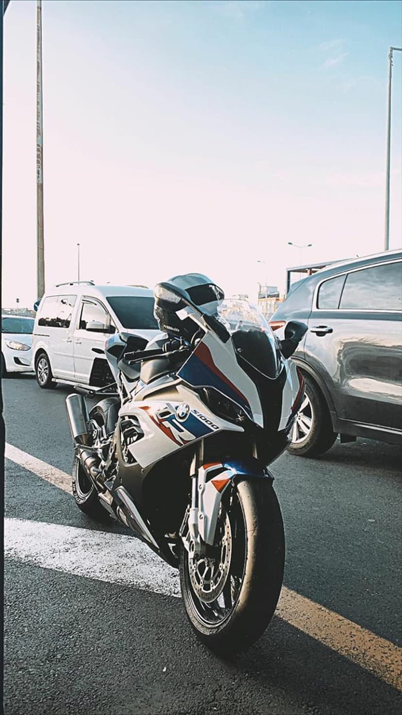 BMW S1000RR sportbike in white, blue, and red parked on asphalt with a van and SUV in blurred traffic background.